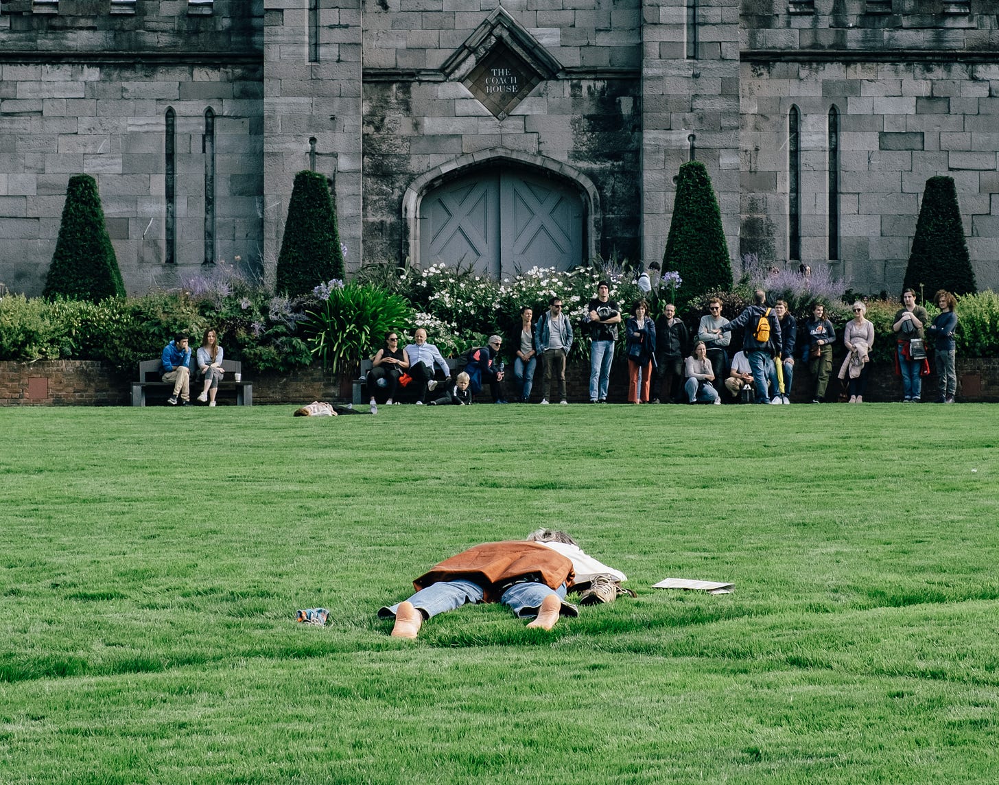 People sitting in a line along a wall in front of a grey stone building with tall gothic windows, while one person lies alone on the grass in the foreground. People sitting in a line along a wall in front of a grey stone building with tall gothic windows, while one person lies alone on the grass in the foreground.
