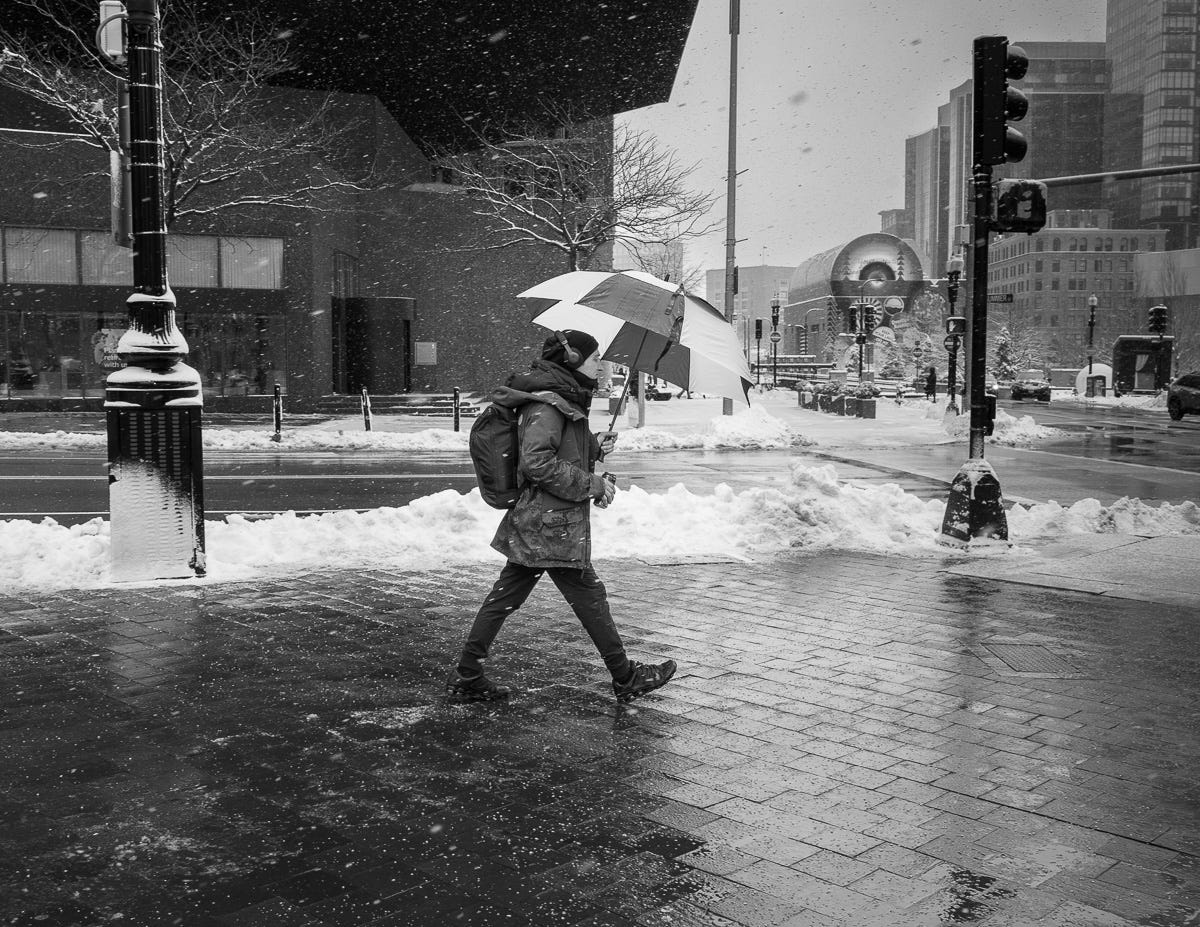 Person walking briskly across a snowy Boston street under an umbrella, framed by icy sidewalks and modern architecture. Person walking briskly across a snowy Boston street under an umbrella, framed by icy sidewalks and modern architecture.