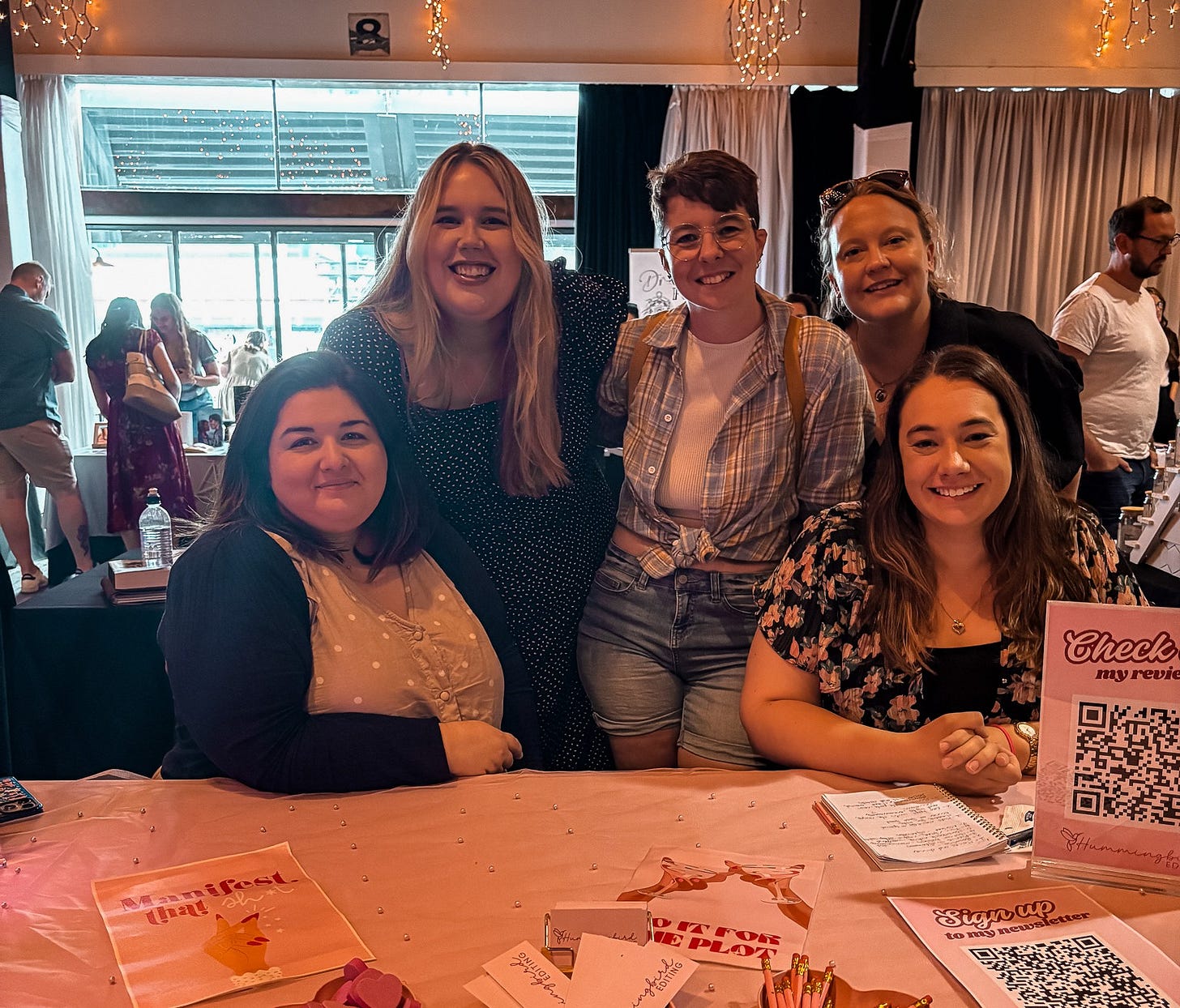 Five people in a photo standing and sitting behind a vendor stall. The stall belongs to Hummingbird Editing. The people behind the stall from left to right are: Laura Ibic (Hummingbird Editing), MG Hunt, Laura Catherine, Krystal from Aussie Indie Reads, and SK May. Five people in a photo standing and sitting behind a vendor stall. The stall belongs to Hummingbird Editing. The people behind the stall from left to right are: Laura Ibic (Hummingbird Editing), MG Hunt, Laura Catherine, Krystal from Aussie Indie Reads, and SK May.