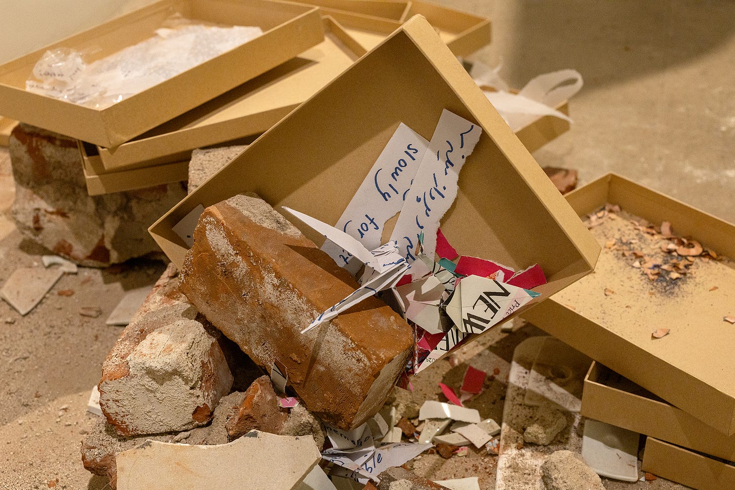 A kraft archive box lies tipped among a pile of reclaimed bricks. A single brick rests inside the box, on top of folded paper cranes and printed fragments.