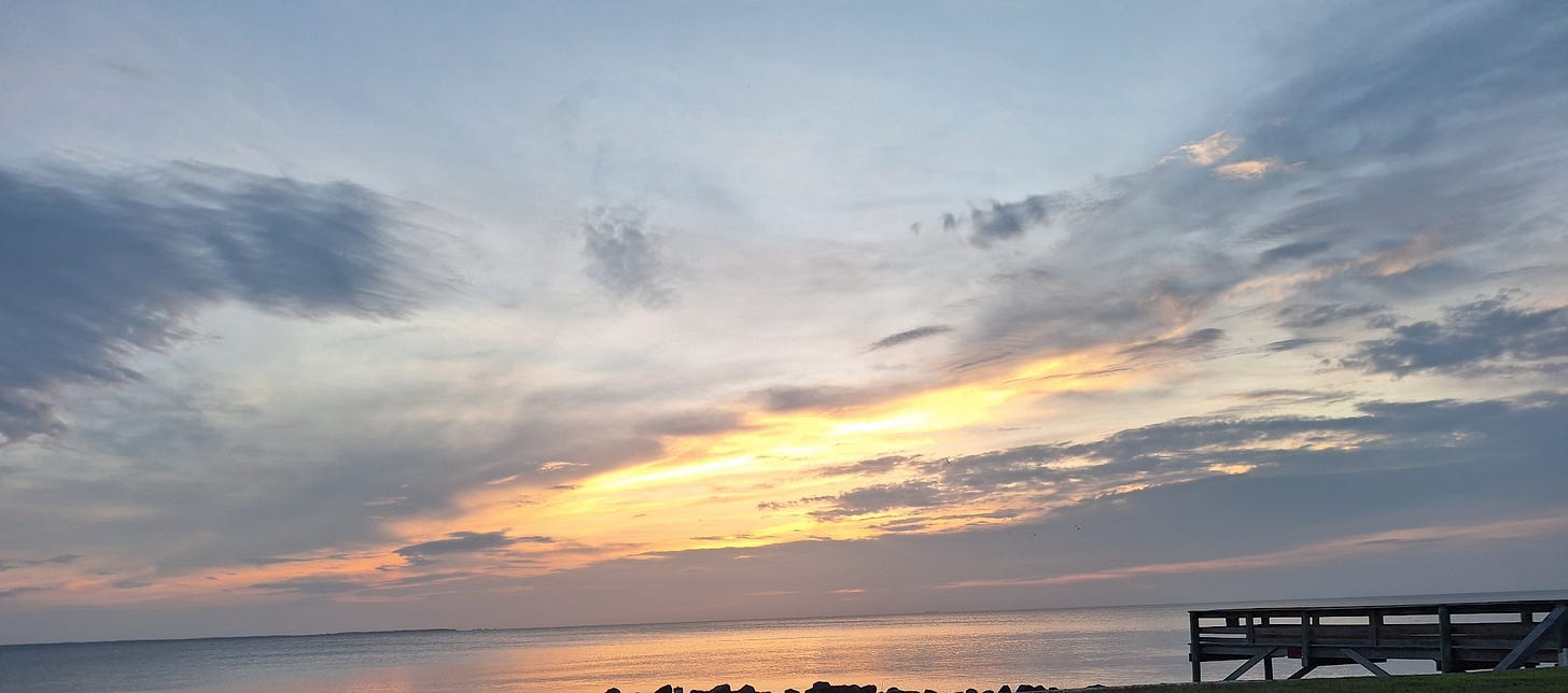 Calm water at dawn with a soft band of light across the horizon, clouds drifting overhead, and a quiet pier along the shoreline.