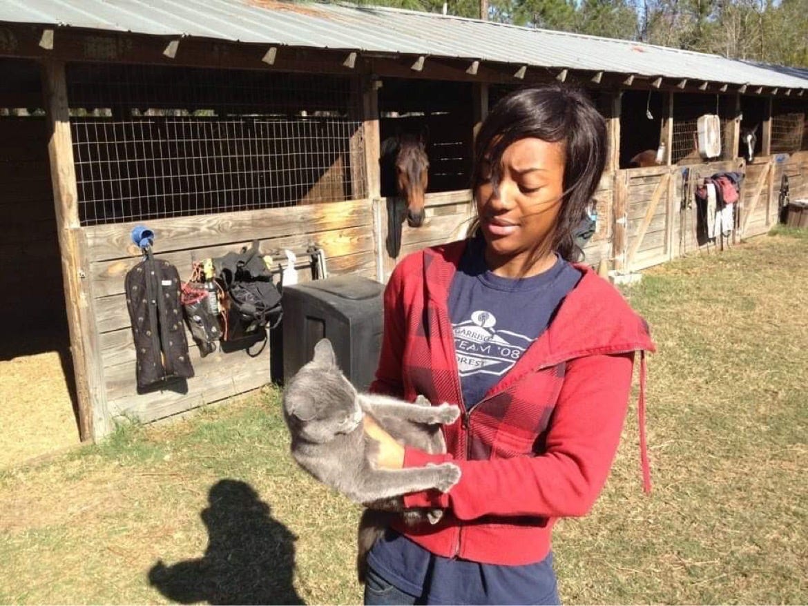 A photo of Kat Lewis at 17 years old. She is on a farm and holding a very angry barn cat. Her facial experience is just as scared and uncertain as the cat's. Several horses are poking their heads out of the stalls behind her.