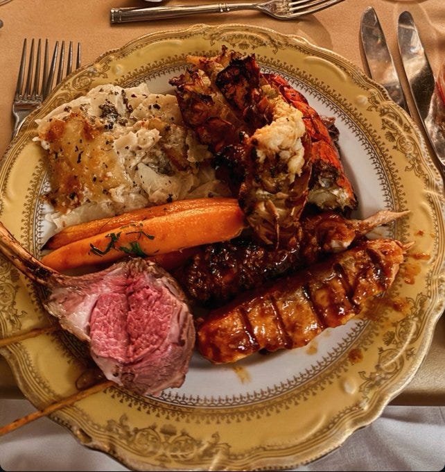 A decorative yellow-rimmed plate holds a lavish meal with pink roasted rack of lamb, grilled steak slices, buttered mashed potatoes garnished with herbs, whole carrots, split lobster tail with drawn butter, and shrimp skewers on sticks, arranged on a beige tablecloth with silver utensils nearby.