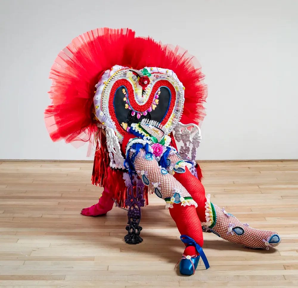 At first, it looks like a slouching body with huge smile as a head. But maybe the head is a butt and we’re behind it. The sculpture is built on a heavy-looking metal chair with floral frills. Everything is ornate with colorful sewn tassles and hems. Bright red tulle creates a crown around a heart-shaped pillow with stuffed doll legs that are 3 or 4 feet long.