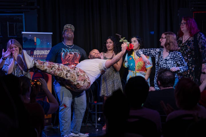 In left photo, a man in a MAGIC THE GATHERING T-shirt and blues jeans holds up another man as several of The Rose improv trouple members look on with a mixture of admiration and surprise. The men being hold is triumphantly holding up a rose.