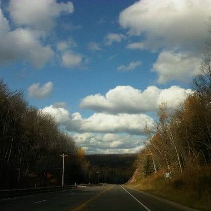 A stretch of road goes into fall trees and bright blue skies with white clouds A stretch of road goes into fall trees and bright blue skies with white clouds