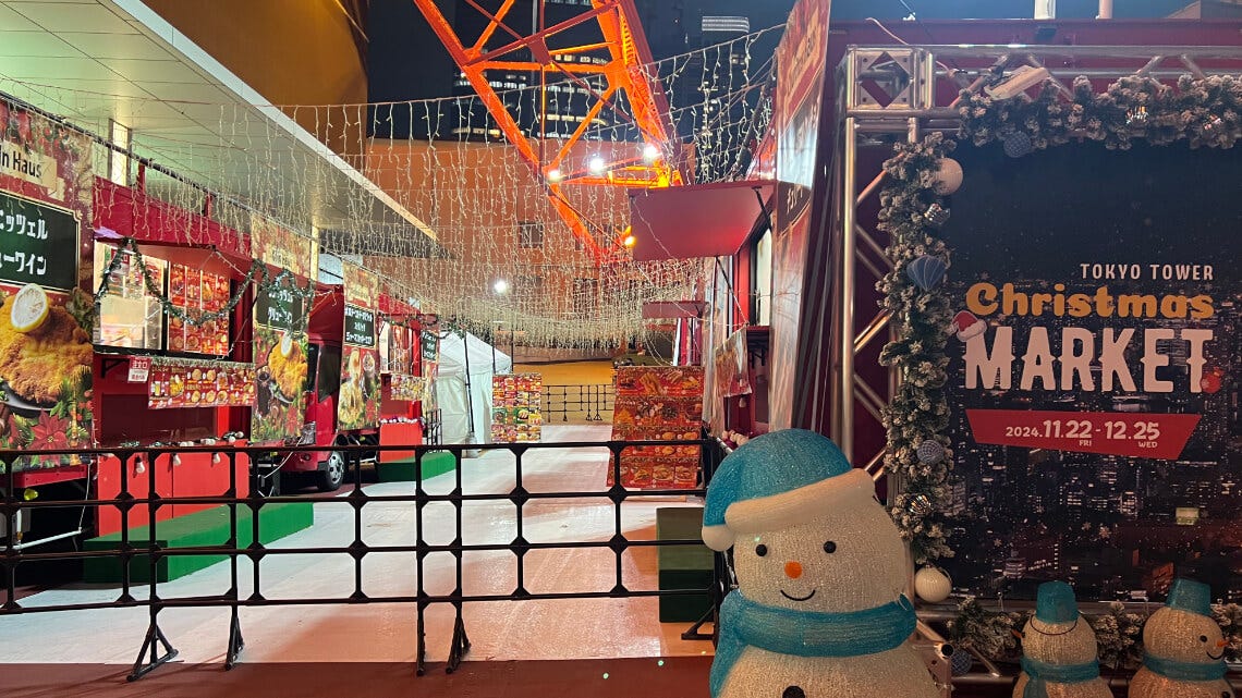 Festive scene at the Tokyo Tower Christmas Market, featuring illuminated food stalls decorated with garlands and lights. A cheerful snowman statue wearing a blue hat and scarf stands in the foreground beside a sign showing the event dates, while strings of warm fairy lights hang overhead under the orange tower structure.