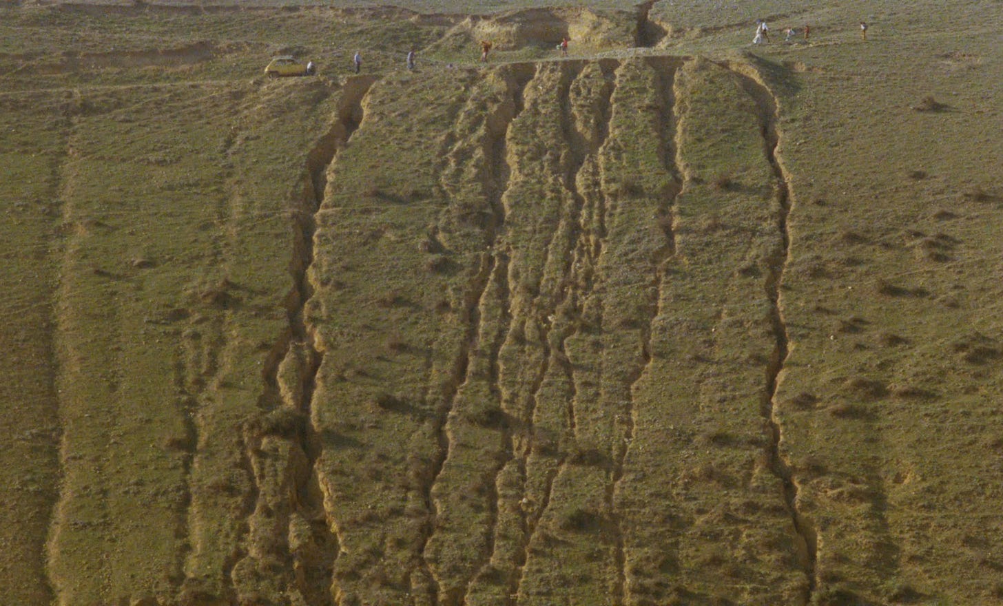 captura de tela do filme "e a vida continua", de abbas kiarostami. é uma tomada aérea mostrando uma estrada que sobe a montanha. a estrada está quase no topo do quadro, com a montanha abaixo. a montanha está cheia de fissuras causadas pelo terremoto. na estrada, várias pessoas caminham carregando objetos e o diretor observa elas de fora do carro, que está parado na frente da maior fissura.