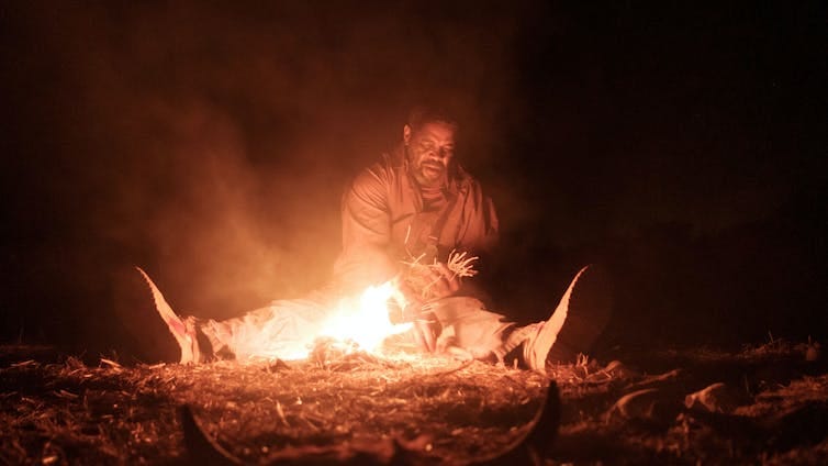 An elderly man sits at a fire, surrounded by darkness.