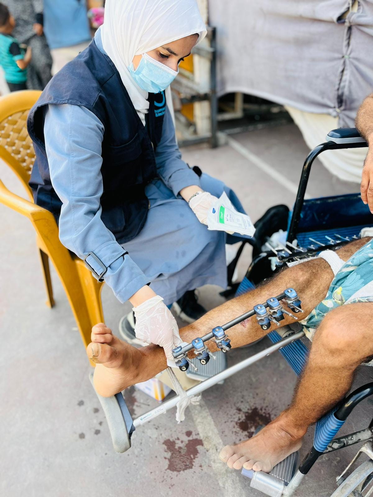 A female healthcare worker wearing a hijab, face mask, and gloves is tending to a man's leg injury while he sits in a wheelchair. The man has an external fixator device attached to his lower leg. The worker is cleaning or dressing the area, using medical supplies. This is taking place outdoors