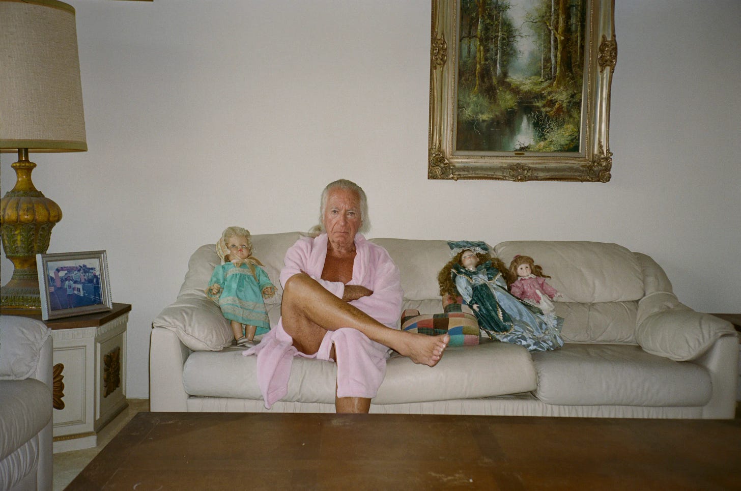 A middle-aged man sits on a beige couch in a sparsely decorated living room, wearing a pink robe and staring directly at the camera, flanked by several dolls placed beside him.