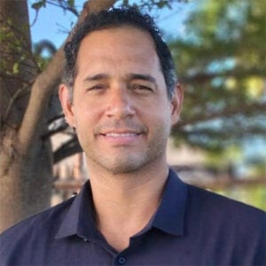 Homem branco, cabelos curtos encaracolados, sorrindo para a foto. Usa camisa polo azul em um ambiente com árvores ao fundo.