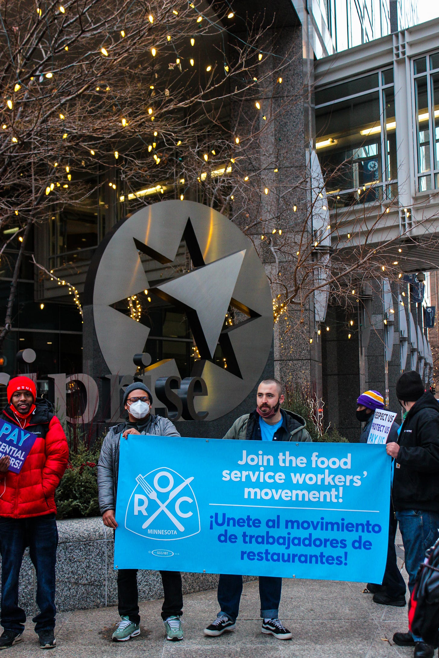 A few crowd members hold a blue sign reading “Join the food service workers’ movement!” A few crowd members hold a blue sign reading “Join the food service workers’ movement!”
