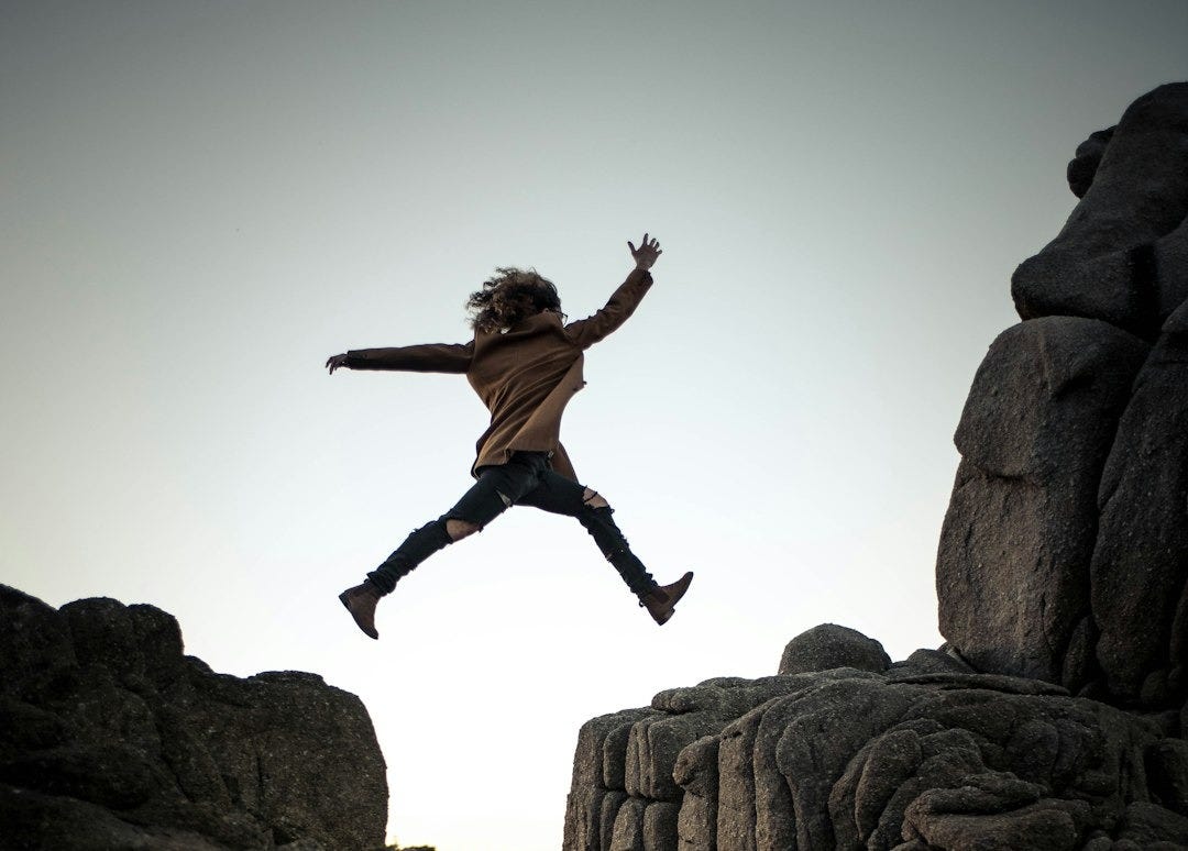 person jumping on big rock under gray and white sky during daytime person jumping on big rock under gray and white sky during daytime