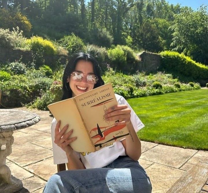 This may contain: a woman sitting on a bench holding a book in her hands and smiling at the camera