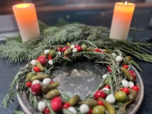 Rosemary Antipasto Wreath - A ring of rosemary on a silver tray with olives, mozzarella balls, and pickles decorating. Two candles and pine fronds in the back.