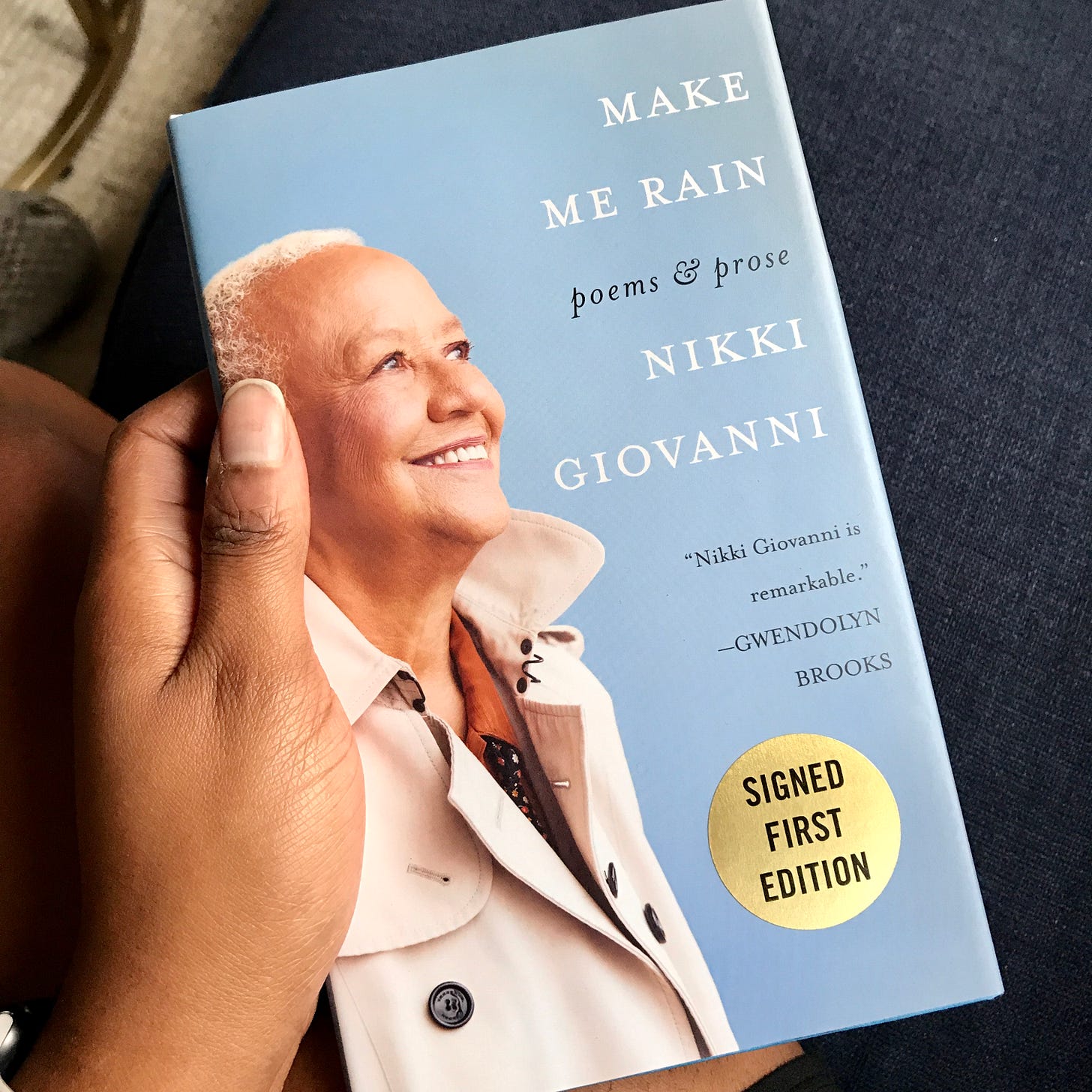 Picture of a Black woman holding a copy of Nikki Giovanni's book, Make Me Rain (Poems & Prose)