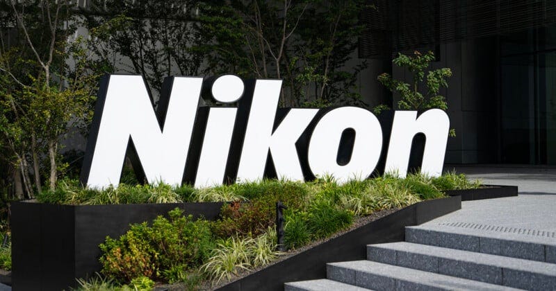 Large, white "Nikon" sign stands amid greenery outside a modern building, near concrete steps and surrounded by trees and bushes.