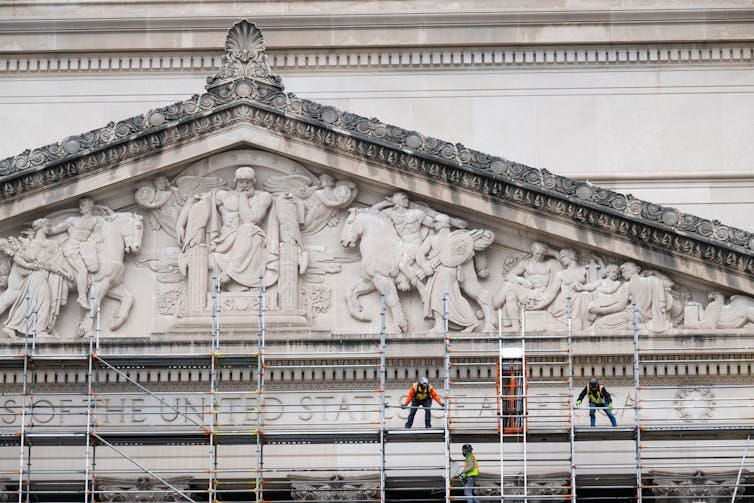 Workers stand on metal scaffolding near a very large, ornate marble building. Workers stand on metal scaffolding near a very large, ornate marble building.