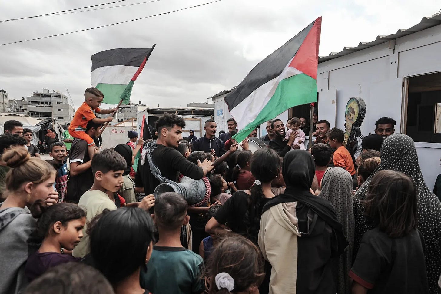 Dozens of people are gathered in a circle. A child in orange is flying one of two Palestinian flags. A man plays a drum.