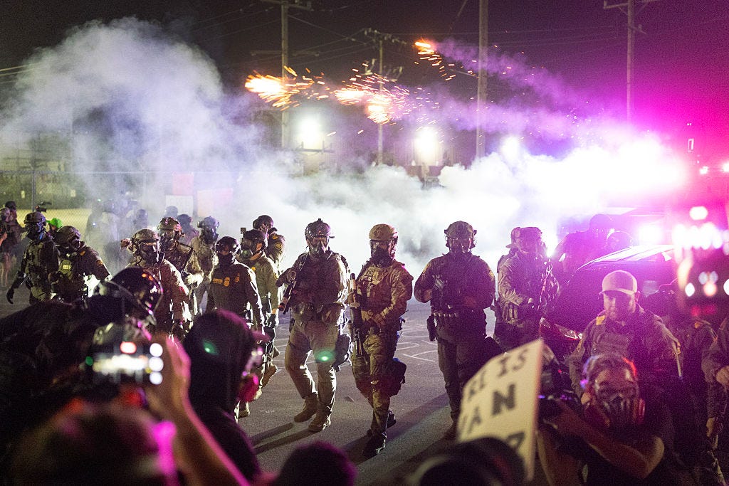 This is a chaotic nighttime scene of a confrontation. A line of heavily armored tactical officers in gas masks advances through thick smoke or tear gas. In the background, fireworks or a similar pyrotechnic device explodes, while the scene is lit by the purple and white lights of an emergency vehicle. In the foreground, civilians, some wearing respirators and holding phones, watch the advance.