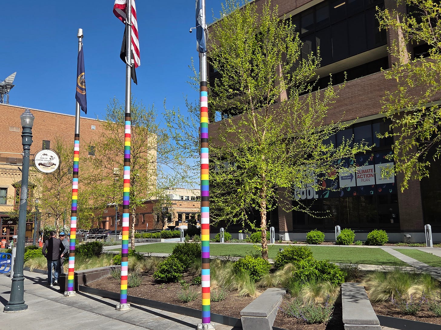 photo of three flagpoles in front of Boise City Hall on a sunny spring day. The flagpoles are wrapped in multi-colored stripes (rainbow, pink, white, light blue, black, and brown) of the Pride/Progress flag. 