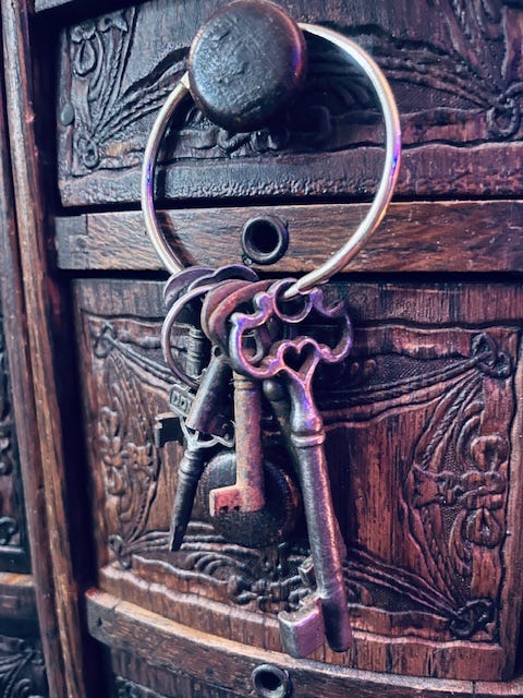 A set of old metal keys hangs from a ring attached to a dark, ornately carved wooden drawer. The keys vary in shape and size, with worn surfaces suggesting age and frequent use.