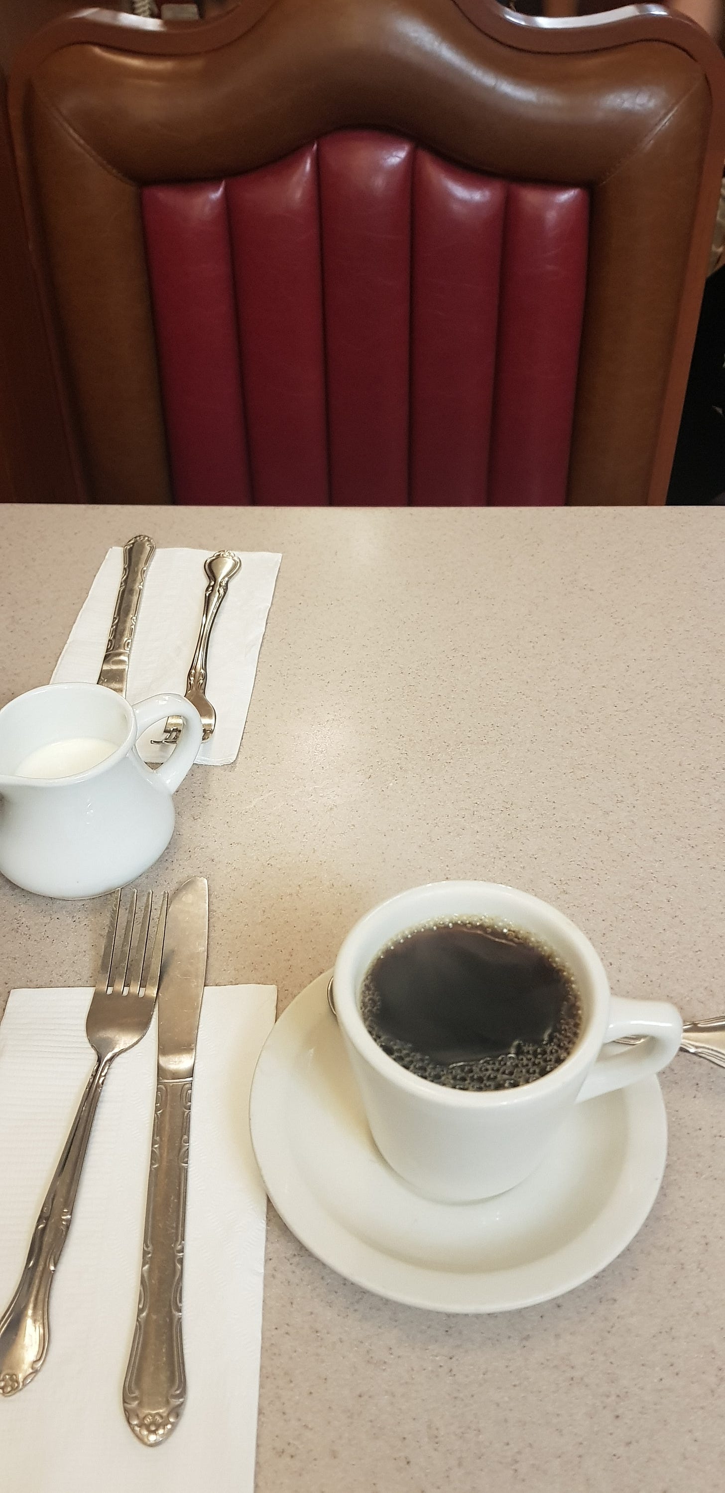 餐館紅色皮質卡座,桌上一杯黑咖啡與銀色餐具,座位空著。 餐館紅色皮質卡座,桌上一杯黑咖啡與銀色餐具,座位空著。