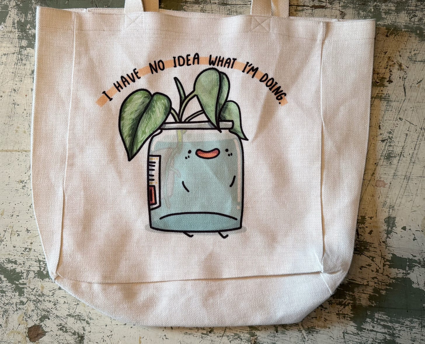 An illustration on a resusable shopping bag showing what appears to be an avocado pit sprouting in a jar of water. The jar has a smiling face, stick arms, and stick legs. Above, the words "I have no idea what I'm doing" arches over the sprouting plant.