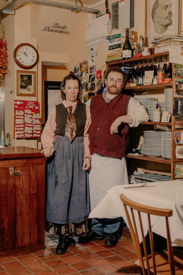 Dos personas vestidas con ropas de colores, una de ellas con una corbata extragrande, están de pie en el comedor de un restaurante abarrotado de estanterías con platos, botellas y otros objetos.