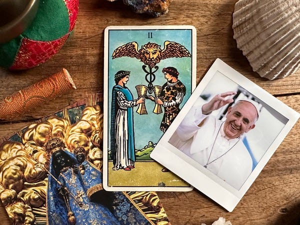 A tarot card with two people on the front with a photo of a man wearing a priest hat and clothing. He is smiling and waving. A bell, a crystal and a photo of the Black Madonna surround the two items.