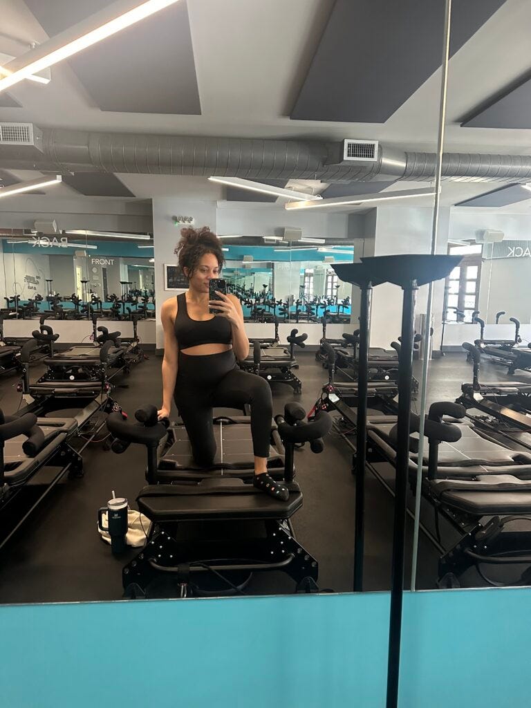 Woman wearing a black sports bra and black high waisted leggings posing on a pilates reformer machine. 