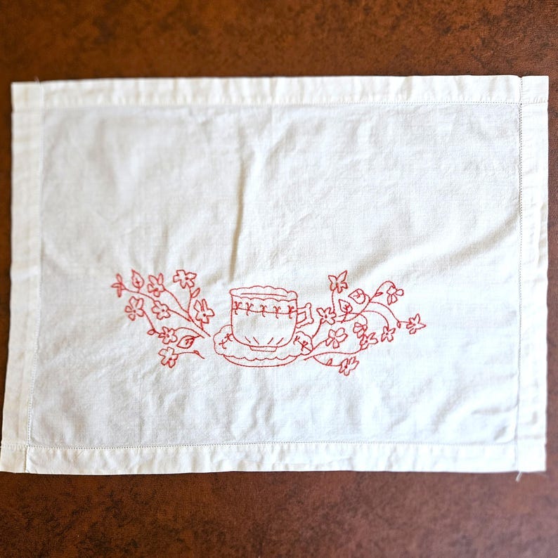 May include: White rectangular placemat with a red embroidered design of a teacup and floral elements. The placemat has a stitched border and is made of a soft, likely cotton, fabric. The embroidery adds a vintage, handcrafted feel.