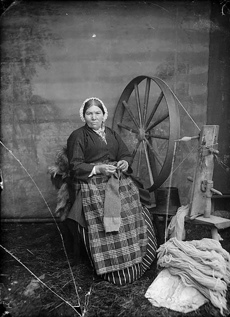 Black and white photograph of a woman in traditional Welsh dress seated beside a large spinning wheel, raw wool on the floor beside her.
