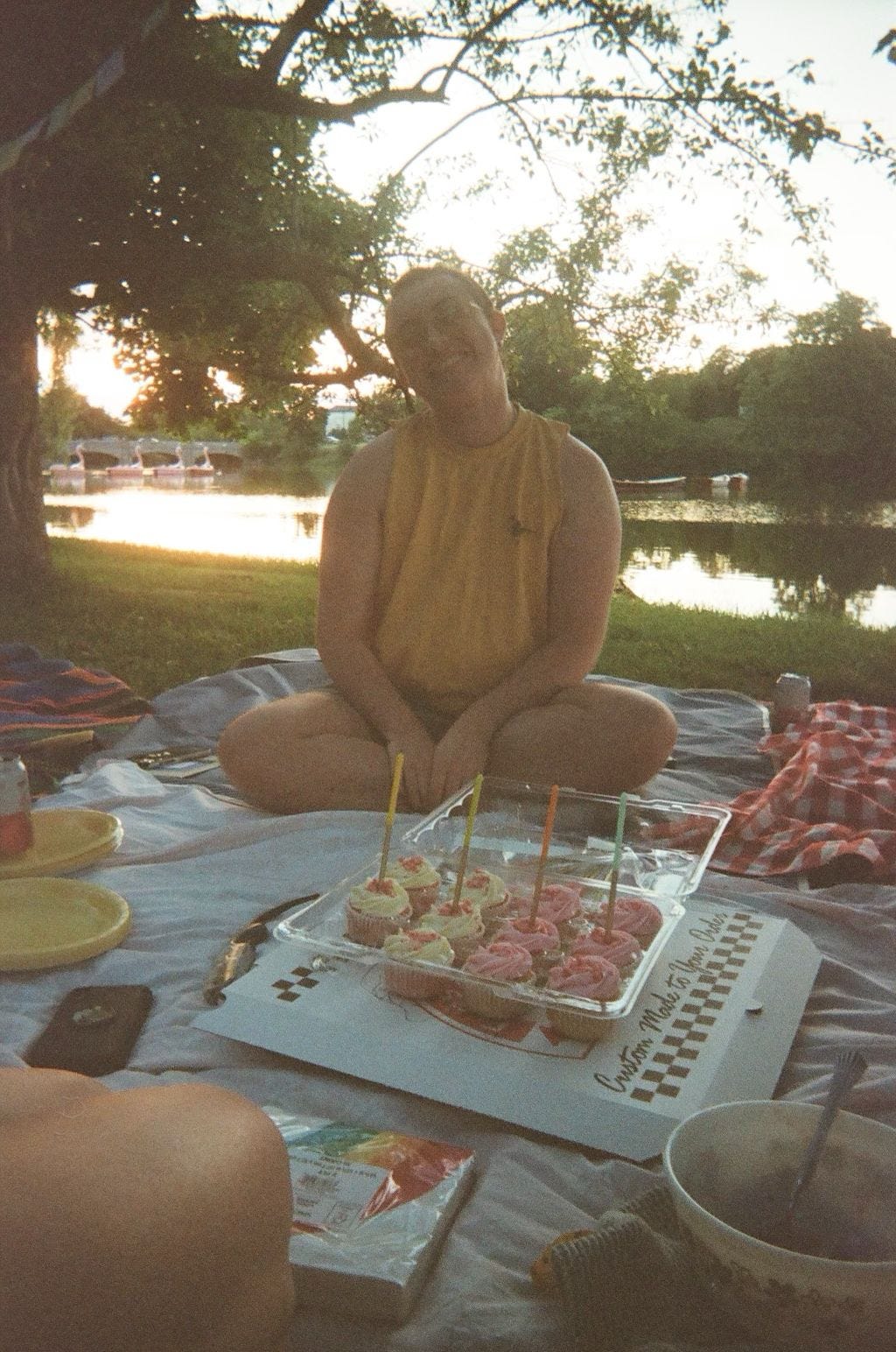 Emory on a picnic blanket with a lake behind him, smiling for the camera with strawberry lemonade cupcakes in front of him