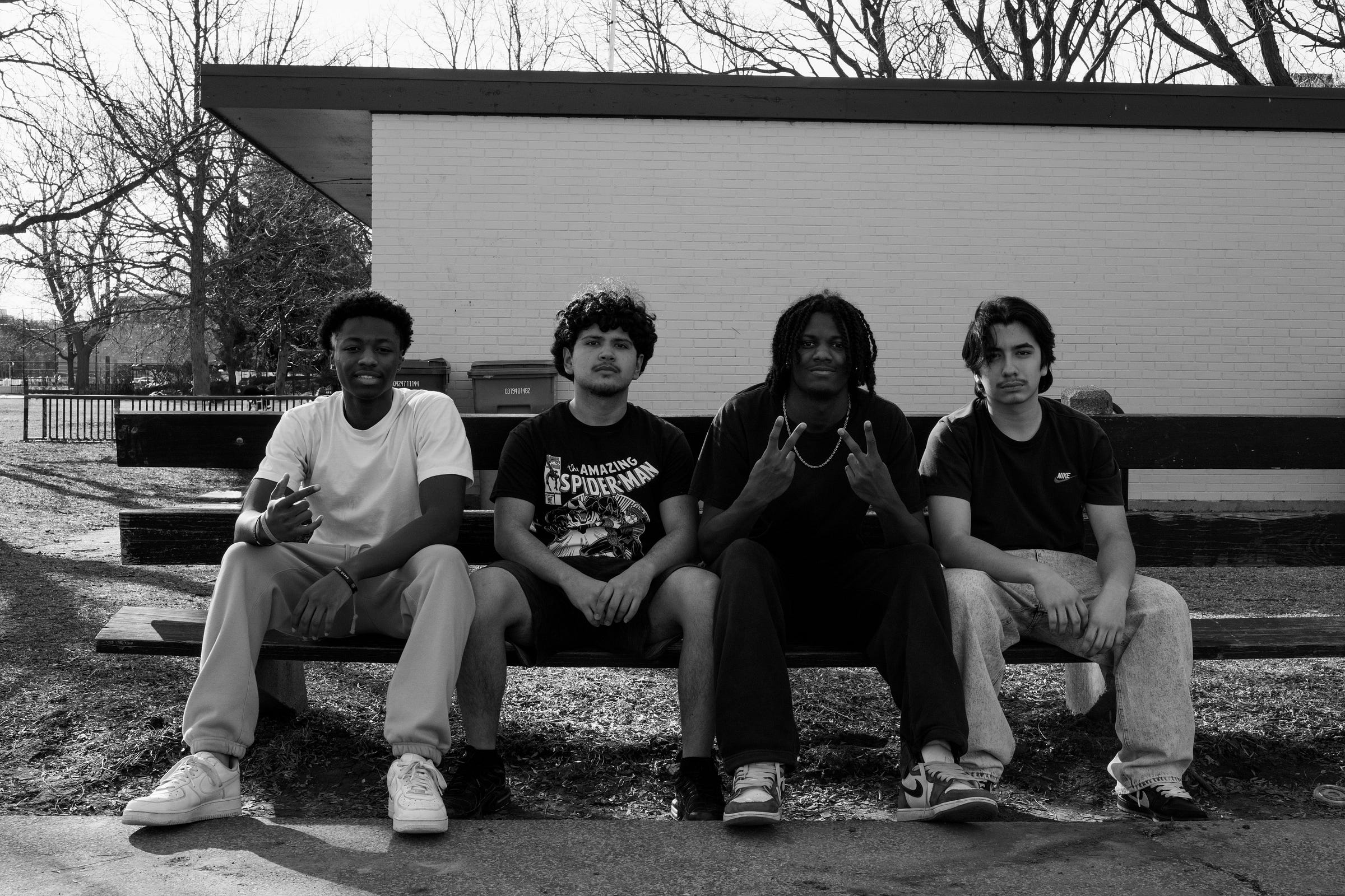 A black-and-white photograph of four young men sitting on a park bench. They face the camera with relaxed but confident expressions, each displaying a unique posture. One wears a Spider-Man t-shirt, another flashes a peace sign, while the others sit with their hands resting on their legs. Behind them, a white brick building, bare trees, and a row of garbage bins set the scene, suggesting an urban park environment. Shadows from the afternoon sun stretch across the ground.