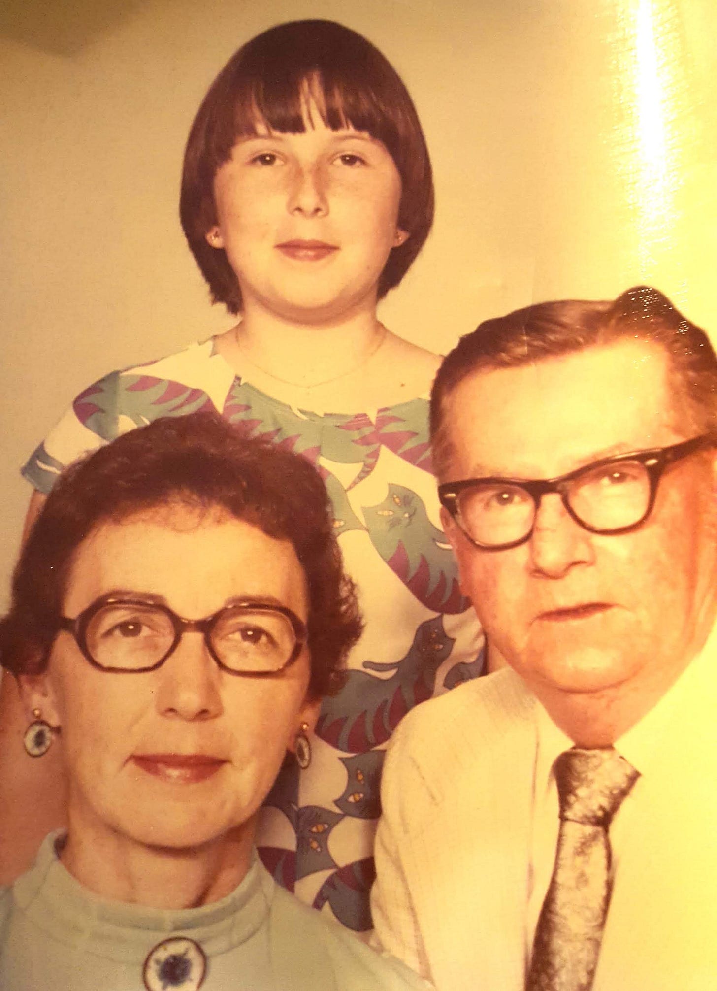 Seventies era family photo. Seated at left: dark haired woman with dark glasses, smiling slightly. To her left, gray haired man with dark glasses, attentive expression. Standing over them on a platform: smiling girl wearing a dress with a purple and blue tabby cat motif. Seventies era family photo. Seated at left: dark haired woman with dark glasses, smiling slightly. To her left, gray haired man with dark glasses, attentive expression. Standing over them on a platform: smiling girl wearing a dress with a purple and blue tabby cat motif.