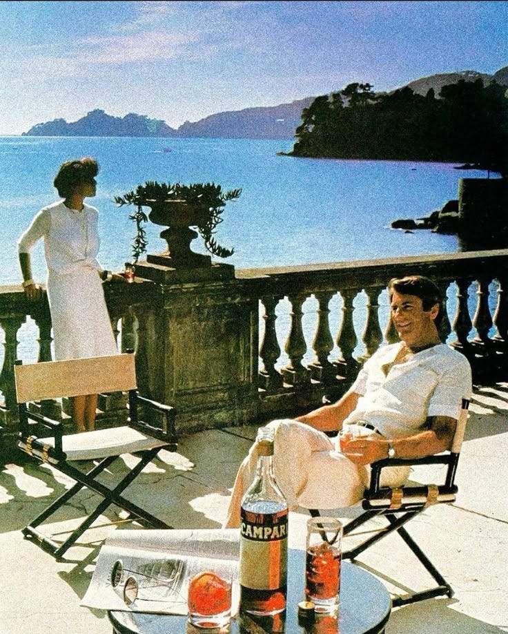 This may contain: two people are sitting on chairs overlooking the water and an outdoor table with drinks in front of them