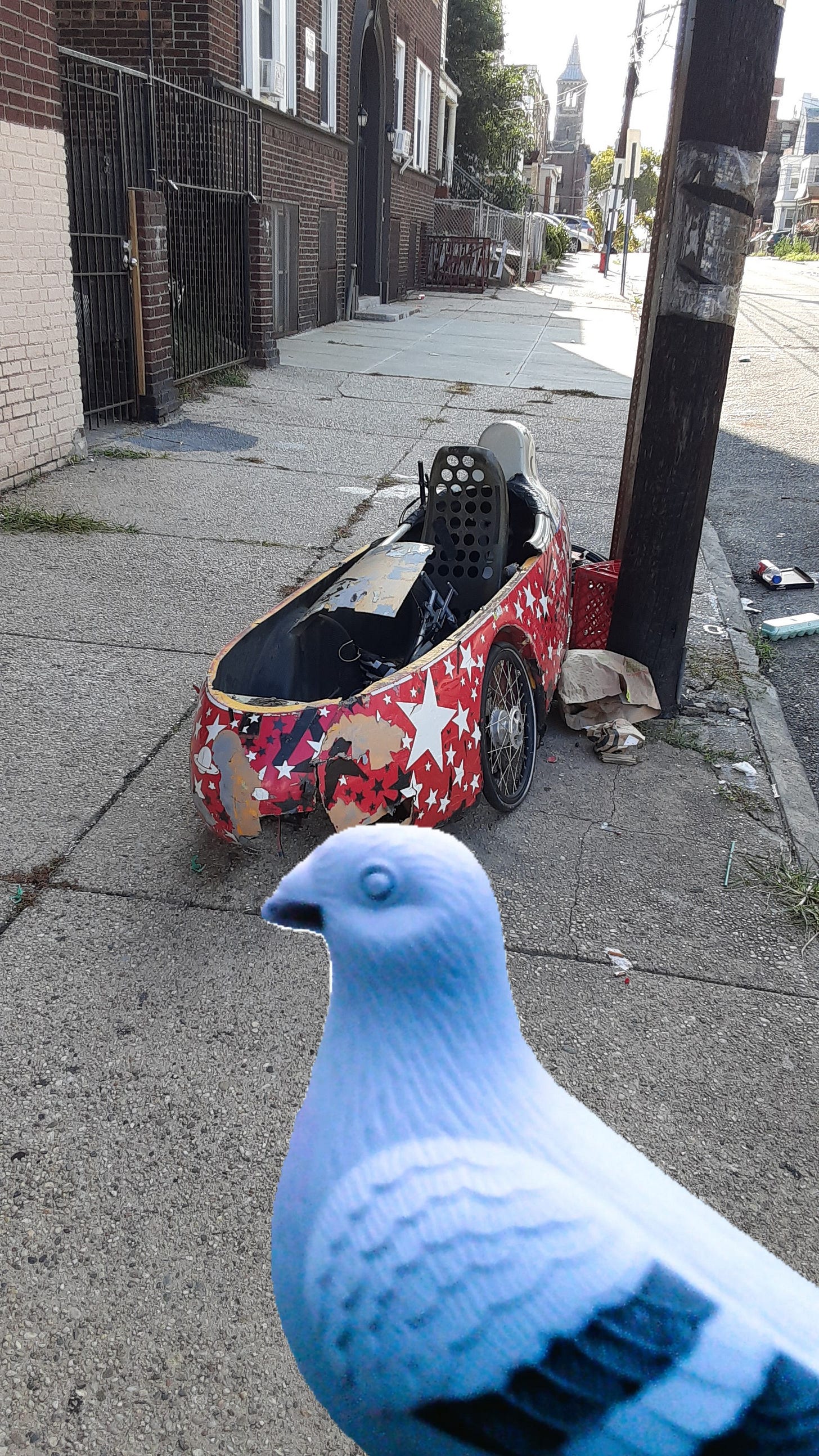 Sidewalk in front of apartment building has a discarded gokart (may be decades old) that has a red color with white stripes. It sits tattered among litter. In foreground is cutout of CB pigeon from another photo. Combined results and both photos by Chilltown Blues (chilltownblues.com)