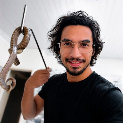 O pesquisador João Miguel, homem branco de cabelo e barba pretos, posa com uma cobra num ambiente interno, provavelmente um laboratório. Traja uma camiseta preta e um óculos de grau com aro prateado. O pesquisador João Miguel, homem branco de cabelo e barba pretos, posa com uma cobra num ambiente interno, provavelmente um laboratório. Traja uma camiseta preta e um óculos de grau com aro prateado.