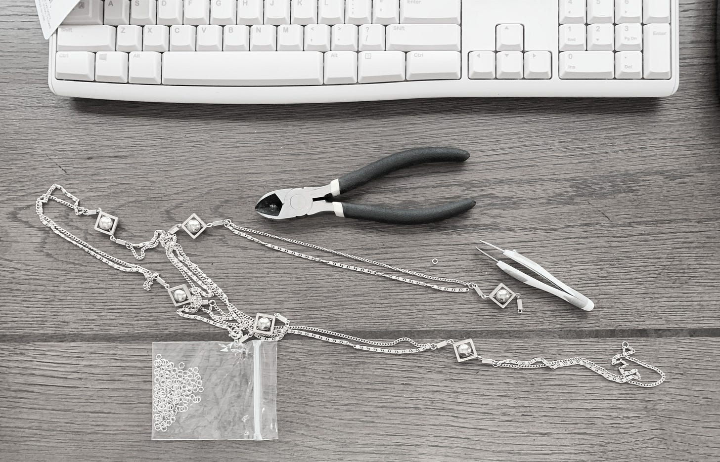 Black and white photo of a desk with a broken necklace, pliers, tweezers, and a package of jump rings Black and white photo of a desk with a broken necklace, pliers, tweezers, and a package of jump rings