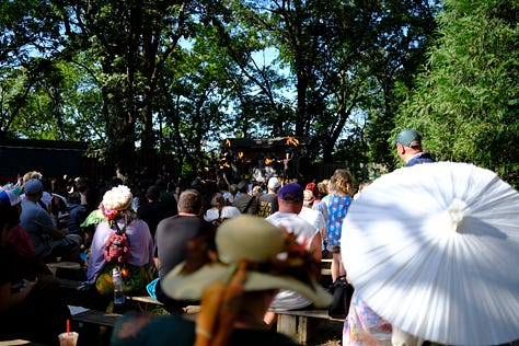 Bards singing in a tavern wearing medieval garb, an elf and wizard surveying goods at a shop, fire-twirler show, Geralt and another warrior deep in conversation, a man playing dead with an arrow under his armpit, guests walking toward the camera, a woman with a horse head looking over a table.