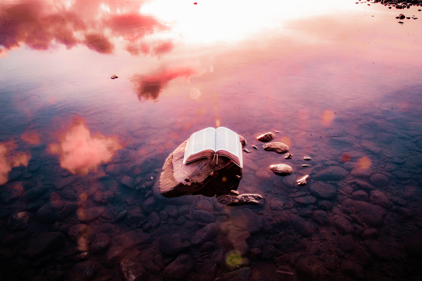 A bible on a rock in a pond with reflections of clouds