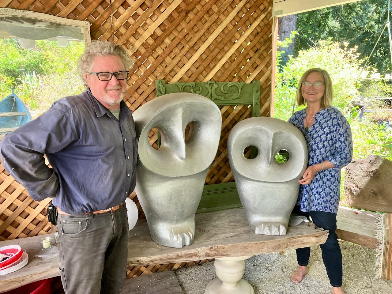 Scott and Ene smiling beside two large owl sculptures they created, displayed on a wooden table in an outdoor studio space.