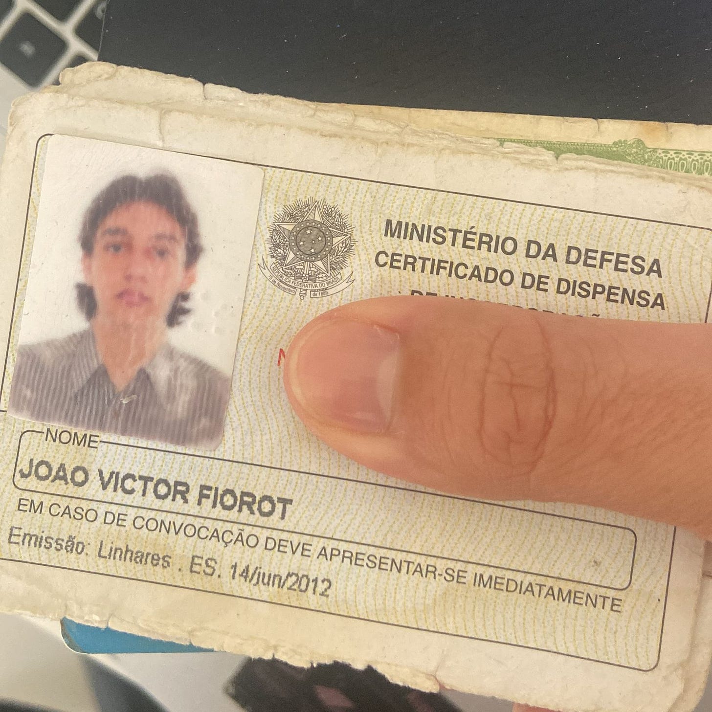 foto do meu certificado de reservista. foto minha antiga, gasta pelo tempo, com uma camisa social listrada, cabelo na altura do pescoço. o documento parece frágil e com todas as suas extremidades já gastadas e manchadas do tempo. um dedo polegar de pele branca cobre o número do documento.