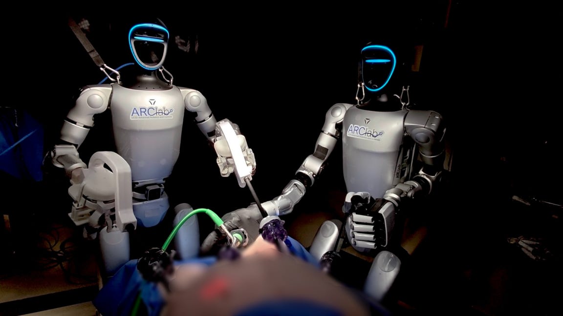 Two humanoid robots stand over a surgical dummy while handling surgical instruments. Two humanoid robots stand over a surgical dummy while handling surgical instruments.