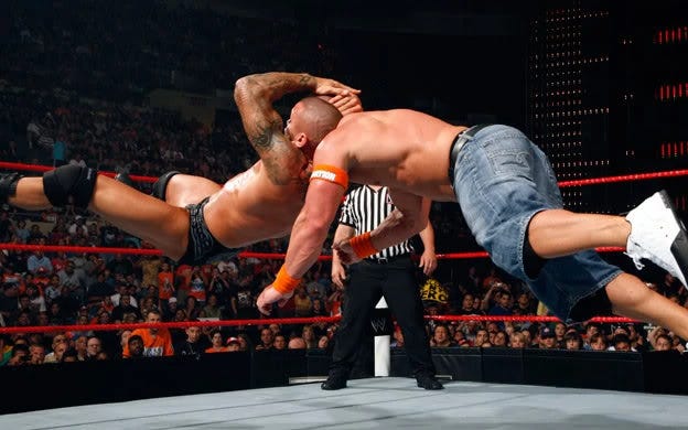 A imagem mostra o lutador profissional Randy Orton a executar o seu movimento de finalização característico, o RKO, em John Cena, durante um combate de wrestling profissional.  Os dois lutadores proeminentes na imagem são Randy Orton (à esquerda, a executar o movimento) e John Cena (à direita, a ser alvo do movimento). O movimento que está a ser executado é o RKO (Randy Keith Orton), um tipo de "cutter" em que o lutador salta na direção do adversário, agarra a sua cabeça numa posição específica e o atira ao chão de cara. A cena ocorre num ringue de wrestling, com um árbitro presente e uma multidão a assistir ao fundo, indicando um evento de luta livre profissional da WWE (World Wrestling Entertainment). 