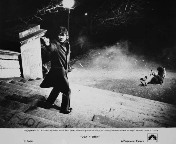 American actor Charles Bronson, wearing a tweed coat, turns to arm a handgun as he stands on a flight of steps, a body lying on the ground at the...