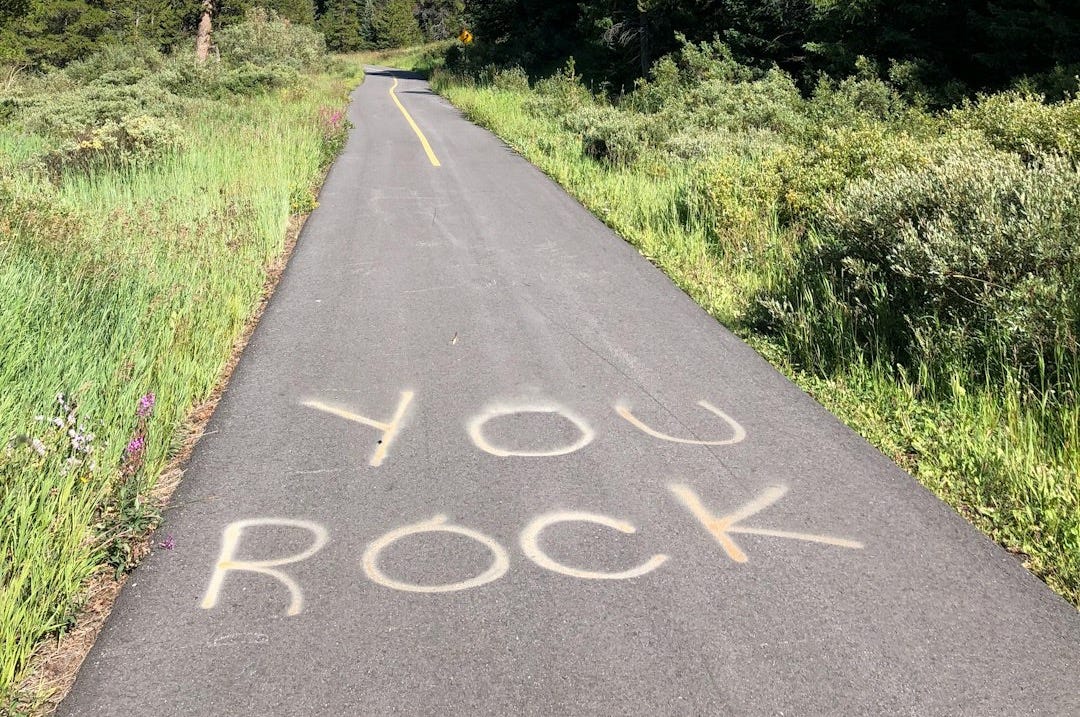gray concrete road with you rock print near green field surrounded with tall and green trees during daytime