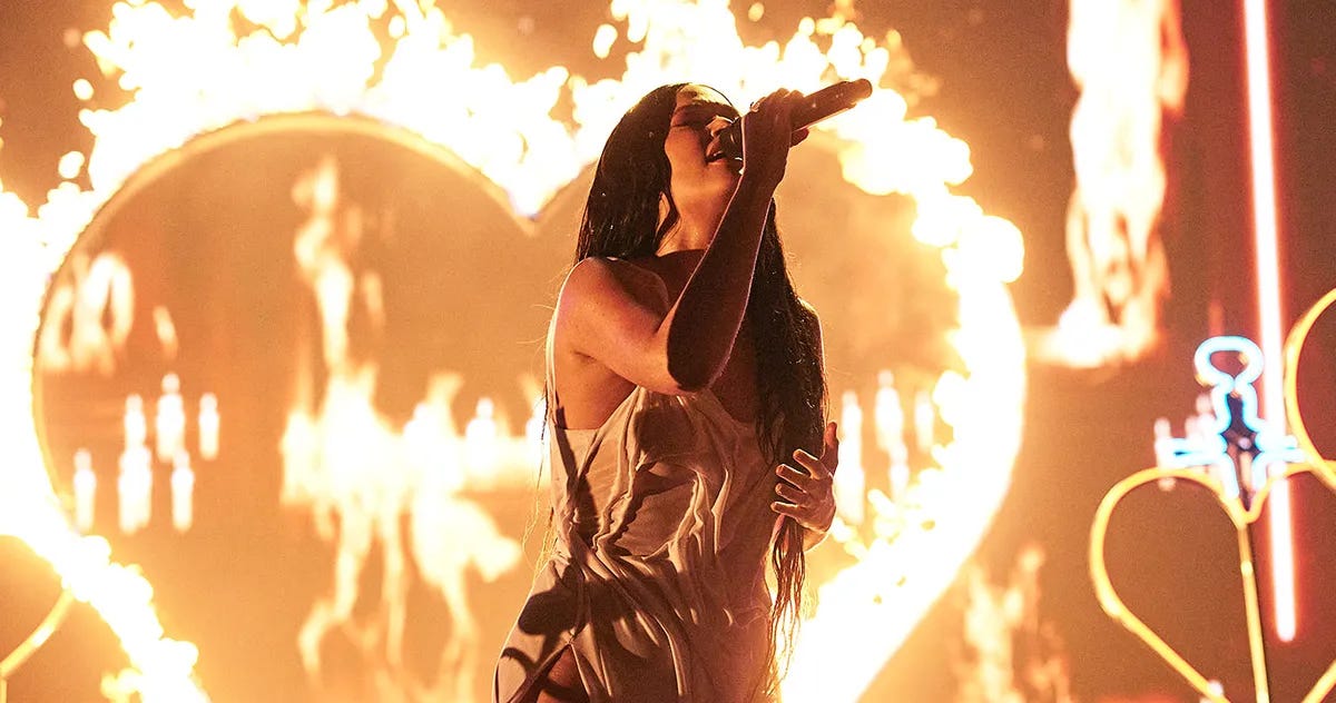 Kacey Musgraves is seen singing into a microphone on stage while a heart-shaped neon sign is set aflame behind her. This renders the entire image in a hue of burnished orange. Kacey Musgraves is seen singing into a microphone on stage while a heart-shaped neon sign is set aflame behind her. This renders the entire image in a hue of burnished orange.
